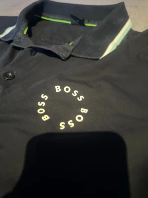 Svart polotopp från BOSS - Säljer en svart polotopp från BOSS med vit logotyp i cirkel på bröstet. Kragen och ärmsluten har vita och ljusgröna ränder som ger en fräsch kontrast. Toppen har korta ärmar och knappar vid halsen. Perfekt för dig som gillar stilrena märkeskläder.