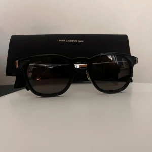 Saint Laurent SL28 svarta solglasögon - Snygga Saint Laurent SL28 solglasögon med svart båge och silvriga detaljer. Modellen har en klassisk rundad form och mörka glas. Metall och acetat ger en lyxig känsla och logga på skalmarna. Perfekta för dig som vill ha en stilren och exklusiv look. Nytt pris 3.500.