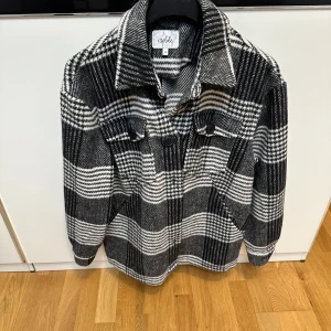 Rutig overshirt i ullmix - Snygg svartvit rutig overshirt med klassisk krage, knäppning framtill och två bröstfickor med knappar. Jackan är i ullmix och har en loose passform, passar bra till höst och vinter.