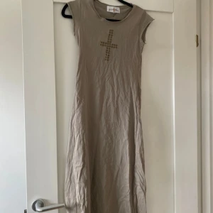 Beige klänning från Ralph & Holly - Beige långklänning från Ralph & Holly med korta ärmar och rund halsringning. Framtill finns ett  kors av små nitar. Klänningen är rak i modellen.
