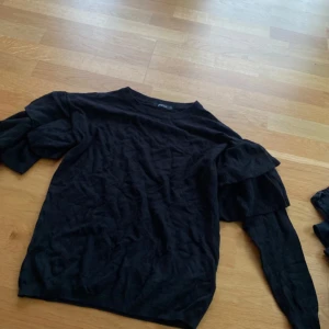 Svart tröja med volangärmar - Svart långärmad tröja med volangdetaljer på ärmarna. Tröjan har en  rund halsringning och har ett mjukt, stickat material.