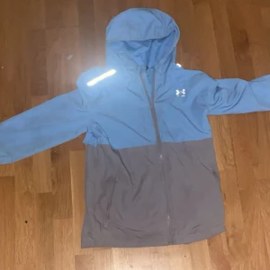 Blå och grå vindjacka från Under Armour - Snygg tvåfärgad vindjacka från Under Armour i blått och grått. Jackan har huva, hel dragkedja framtill och två sidofickor. Perfekt för blåsiga dagar och har en sportig vibe med reflekterande detalj på axeln.