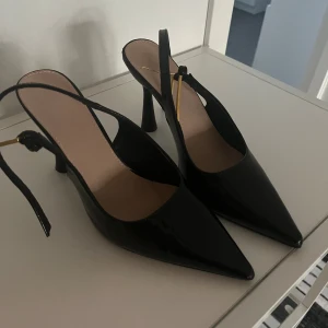 Klackar  - Snygga svarta pumps med spetsig tå och hög klack. Skorna har en elegant rem med gulddetalj runt hälen och är tillverkade i glansigt syntetmaterial. Perfekta för dig som vill ha en stilren och trendig look.