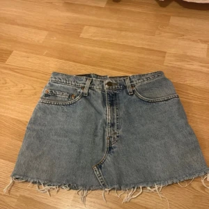 Levi's jeanskjol med fransig kant - Säljer en klassisk blå jeanskjol från Levi's med fransig, rå kant nertill. Kjolen har fem fickor, bälteshällor och knappstängning framtill. Perfekt för dig som gillar en avslappnad och cool stil.