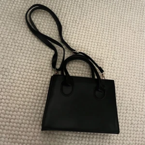 Svart handväska  - Snygg svart handväska med stilren, fyrkantig form och både handtag och lång axelrem. Perfekt storlek för det viktigaste och passar till många olika outfits.