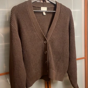 Brun stickad kofta från H&M - Mysig brun stickad kofta från H&M med grov ribbstickning och v-ringning. Koftan har långa ärmar och stängs med fyra knappar framtill. Perfekt att slänga över en t-shirt eller linne för en chill look.