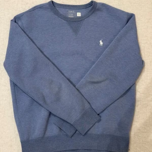 Blå sweatshirt från Polo Ralph Lauren - Snygg blå sweatshirt från Polo Ralph Lauren med klassisk rund halsringning och broderad vit logga på bröstet. Tröjan har långa ärmar och ribbade muddar vid ärmslut och nederkant. 