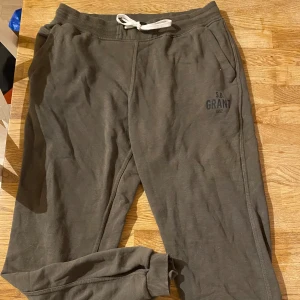 Olivgröna mjukisbyxor S.B. Grant XL - Olivgröna mjukisbyxor från S.B. Grant med vit dragsko i midjan och muddar vid bensluten. Byxorna har fickor på sidorna och tryckt logga på ena benet. Perfekta för chill eller träning.