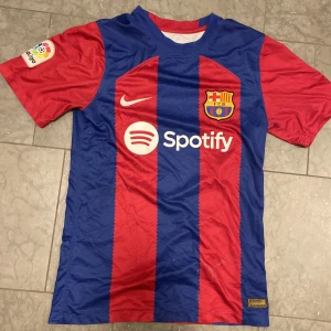 FC Barcelona matchtröja - Säljer en officiell FC Barcelona matchtröja från Nike med klassiska blå och röda vertikala ränder. Tröjan har klubbmärke, La Liga-patch på ärmen och sponsorloggor från Spotify och UNHCR. Tillverkad i lätt och ventilerande material, perfekt för fotbollsfans.