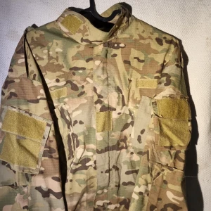 Camouflage kostym med fickor - Säljer en militärinspirerad kostym i klassiskt camouflage-mönster med flera praktiska fickor och kardborrepatchar på armar och bröst. Färgerna går i beige, grön och brun. Materialet är slitstark bomull och polyester, perfekt för dig som gillar utility och streetstyle.