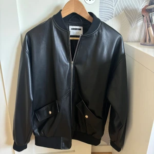 Svart bomberjacka från Noisy May - Snygg svart bomberjacka från Noisy May i skinnimitation. Jackan har dragkedja framtill, två stora fickor med tryckknappar och ribbade muddar vid ärmslut och nederkant. Perfekt för dig som gillar en clean och stilren look.