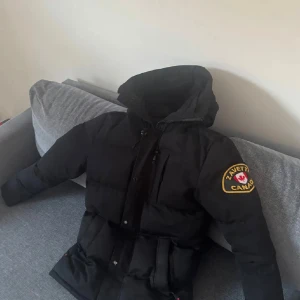 Svart pufferjacka från Zavetti Canada - Svart pufferjacka från Zavetti Canada med stor huva och snygg fuskpäls. Jackan har en tydlig logga på ärmen, dragkedja framtill och flera fickor. Perfekt för kalla dagar med sin tjocka vaddering och stilrena look.