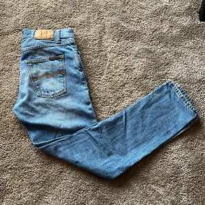 Ljusblå Nudie Jeans Steady Eddie II  - Ljusblå jeans från Nudie Jeans, modell Steady Eddie II All Day Blues. Klassisk design, knappgylf och snygga sömmar. Tillverkade i 100% bomull med Nudie-logga på bakfickan. Perfekt för dig som gillar en avslappnad och tidlös look. Jeansen är i storlek W28 L30. 