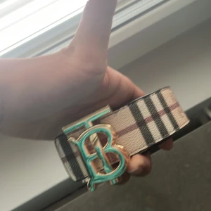 Burberry bälte med rutigt mönster - Säljer ett brett Burberry bälte med klassiskt rutigt mönster i beige, svart och rött. Bältet har en stor guldfärgad spänne med turkos detalj och tydlig TB-logga. Ena sidan är beige med rutor, andra sidan är svart med präglat mönster och logga. Perfekt för att lyfta din outfit.