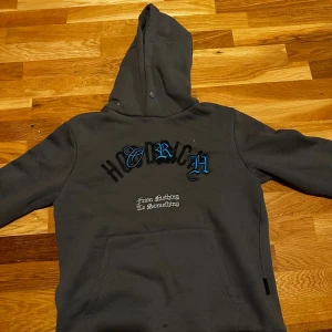 Mörkgrå hoodie från Hoodrich - Säljer en mörkgrå hoodie från Hoodrich med stor huva och tryck både fram och bak. Framsidan har blå och svart text samt broderad text, medan baksidan har stort svart tryck. Klassisk känguruficka och skön passform, perfekt för streetwear-stilen.