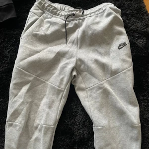 Grå mjukisbyxor från Nike - Snygga grå mjukisbyxor från Nike med svart logga på vänster ben. Byxorna har elastisk midja med snörning och är tillverkade i mjukt bomullsmaterial. Perfekta för chill eller träning, med bekväm passform och praktiska fickor.