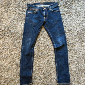 Mörkblå skinny jeans från Nudie - Säljer ett par mörkblå skinny jeans från Nudie med klassiska orange sömmar och vågig brodyr på bakfickorna. Jeansen har fem fickor, låg midja och är tillverkade i stretchig denim för en tight passform. Perfekta för dig som gillar en smal siluett. W30 L32.