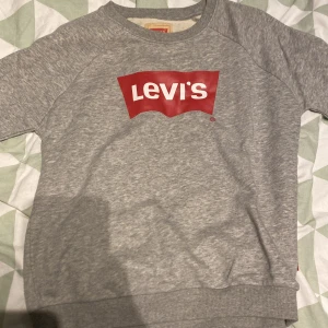 Grå sweatshirt från Levi's med logga - Säljer en grå sweatshirt från Levi's med den klassiska röda loggan framtill. Tröjan har rund halsringning, långa ärmar och ribbade muddar vid ärmslut och nederkant. Perfekt för en chill och stilren look.