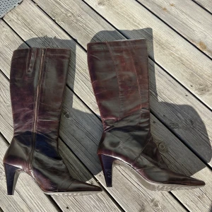 Bruna höga stövlar i skinn med klack - Snygga mörkbruna stövlar i äkta skinn med spetsig tå och hög klack. Stövlarna har dragkedja på insidan och en elegant, slank siluett. Perfekta för dig som vill ha en klassisk och stilren look med lite edge.