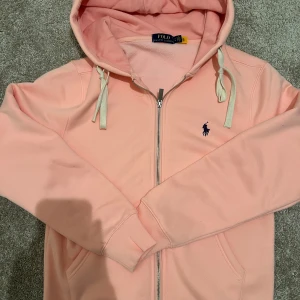 Rosa hoodie från Polo Ralph Lauren - Snygg ljusrosa hoodie från Polo Ralph Lauren med dragkedja och vit snörning i huvan. Riktigt varm och skön till vintern. Även en av de snyggaste tröjorna jag haft. Dock är den helt ny nästan. Använt 5-6 gånger. Inga hål eller nåt. Bara riktigt fet!