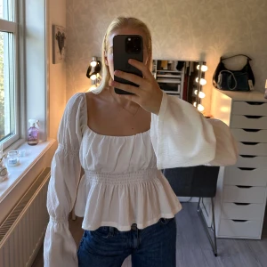 Vit blus med puffärmar och smock - Supersöt vit blus med fyrkantig ringning, puffiga långa ärmar och smockad midja ifrån Ginatricot. Blusen har volang nertill och är tillverkad i ett lätt, strukturerat bomullstyg. Perfekt att styla med jeans för en trendig look. Använd ett fåtal gånger.💖