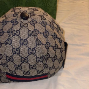 Gucci keps med GG-mönster och röd-blå rand - Snygg Gucci keps med klassiskt GG-mönster i beige och grå toner. Kepsen har en röd och marinblå rand på sidan och justerbar rem i svart skinn baktill. Perfekt för dig som vill ha en lyxig och trendig accessoar med ikonisk design.