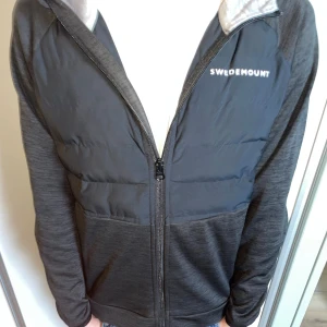 Svart hybridjacka från Swedemount - Snygg svart hybridjacka från Swedemount med vadderad framsida och mjuka ärmar och rygg i stretchigt material. Jackan har huva, hel dragkedja och en stilren look med logga på bröstet. Perfekt för lager-på-lager under kyliga dagar.