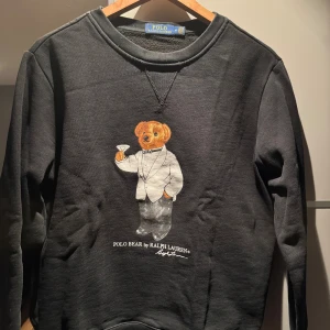Svart sweatshirt Polo Bear Ralph Lauren - Svart sweatshirt från Polo Ralph Lauren med ikoniska Polo Bear-motivet på bröstet. Tröjan har rund halsringning och långa ärmar. Mjuk insida och klassisk passform, perfekt för dig som gillar streetstyle med en twist.