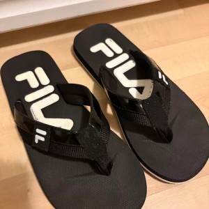 Svarta sandaler från Fila - Svarta sandaler från Fila med breda remmar i syntet och textil. Sulan är platt och har ett vågigt mönster undertill för extra grepp. Stor Fila-logga i vitt på fotbädden ger en sportig vibe. Perfekta för sommaren och chill dagar.