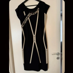 Svart kortklänning med pärldetaljer - Snygg svart kortklänning med beige dragkedjedetaljer och pärlor längs ena axeln. Klänningen har en modern design med korta ärmar och rund hals. Perfekt för dig som gillar statement-plagg med coola detaljer.