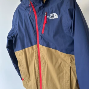Blå och beige vindjacka från The North Face - Snygg tvåfärgad vindjacka från The North Face i marinblått och beige med röda dragkedjor. Jackan har huva, bröstficka med dragkedja och logga på bröstet och ryggen. Tillverkad i slitstark polyester, perfekt för blåsiga dagar. Använd ett fåtal gånger och syns inte att den varit använd. Inga skador, superfin i kulörerna.