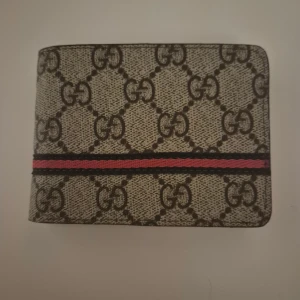  Plånbok med GG-mönster och röd rand - Snygg plånboki med klassiskt GG-mönster i beige och brunt. Utsidan har en röd och svart rand som ger en extra touch. Insidan är mörkbrun med plats för kort och sedlar. Perfekt accessoar för dig som gillar lyxiga detaljer.