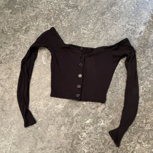 Svart croppad ribbad bolerotopp - Säljer en svart croppad bolerotopp med långa ärmar och knappar framtill. Toppen är ribbad och har en offshoulder-design som ger en snygg och trendig look. Perfekt att styla med höga jeans eller kjol.