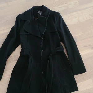 Svart kappa med bälte från Suxe - Stilren svart kappa från Suxe med klassisk krage, knappar framtill och bälte i midjan. Jackan har långa ärmar och markerade muddar med knappar. Perfekt för dig som gillar en tidlös och elegant look.