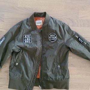 Olivgrön bomberjacka från KappAhl - Snygg olivgrön bomberjacka från KappAhl med coola NYC-patchar och orange foder. Jackan har dragkedja framtill, ribbade muddar och fickor med tryckknappar. Perfekt för dig som gillar streetstyle och vill sticka ut lite extra.