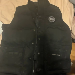 Svart dunväst från Canada Goose XS - Säljer en svart dunväst från Canada Goose i storlek XS. Västen har hög krage, dragkedja och knappar framtill samt två stora fickor. Klassisk logga på bröstet och diskret Canada Goose-märke på fickan. Perfekt för kyliga dagar.