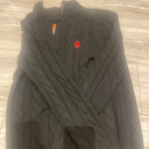 Svart kabelstickad tröja Polo Ralph Lauren - Svart kabelstickad tröja från Polo Ralph Lauren med halv dragkedja och röd broderad logga på bröstet. Tröjan har ribbade muddar och hög krage, perfekt för kyliga dagar. Klassisk och stilren design som funkar till allt. Knappt använt