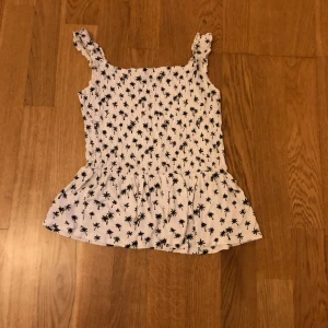 Vit smockad linnetopp med palmer H&M 158-164 - Söt vit linnetopp från H&M med svarta palmtryck. Toppen har smockad överdel, volang nertill och breda axelband med rynkade detaljer. Perfekt för sommarens varma dagar och ger en chill vibe. Är sydda på banden syns inte jätte mycket men har lite rosa tråd på ena bandet.