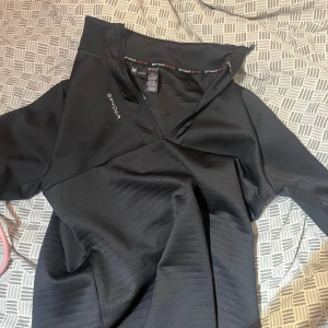 Svart sporttröja från Spyder - Säljer en svart sportig tröja från Spyder med halv dragkedja och diskret logga på bröstet. Tröjan har lång ärm och är tillverkad i ett stretchigt syntetmaterial som passar perfekt för träning eller aktiva dagar. Helt ny