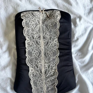 Snygg korsett - Svart korsett från Closeness i storlek S. Korsetten har en bred, dekorativ spets i beige framtill och en liten rosett upptill. Modellen är axelbandslös och kort, med figurnära passform och hakknäppning på insidan. Perfekt för dig som gillar detaljer och en chic look.