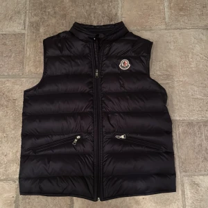 Svart dunväst från Moncler - Svart dunväst från Moncler med klassisk quiltad design och hög krage. Västen har två snedställda fickor med dragkedja och Moncler-logga på bröstet. Perfekt för lager-på-lager och snygg till både hoodie och t-shirt. Storlek 164 cm / 14 år köpt på NK
