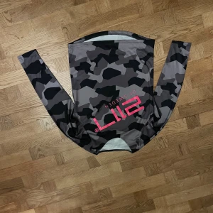 2117 of Sweden camo MTB tröja - Säljer en långärmad sporttröja från 2117 of Sweden med grått och svart camouflagemönster och rosa logga på bröstet. Tröjan är tillverkad i polyester och har en tight passform, perfekt för träning eller utomhusaktiviteter. Spelfilm för MTB