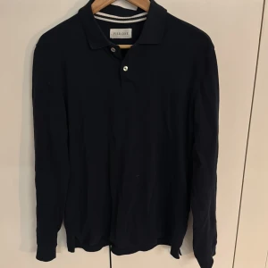 Mörkblå långärmad pikétröja Pier One - Säljer en stilren mörkblå långärmad pikétröja från Pier One. Tröjan har klassisk krage, knappar vid halsen och är tillverkad i mjuk bomull. Perfekt för dig som gillar en clean och enkel look.