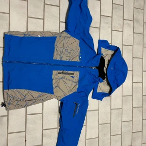 Blå vindjacka Peak Performance Junior - Snygg blå vindjacka från Peak Performance Junior med gråa partier och grafiskt mönster. Jackan har huva, dragkedja framtill och praktiska fickor. Perfekt för kalla dagar och outdoor-aktiviteter. Materialet är slitstarkt och jackan har coola detaljer som tryckknappar och zip på ärmen. Och finns inget mer såna här som säljs så den är antik och värdefull!