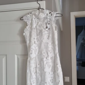 Vit spetsklänning från Gina Tricot - Superfin vit kort spetsklänning från Gina Tricot med blommigt mönster och hög hals. Klänningen är ärmlös och har dragkedja i ryggen. Perfekt för student eller sommarens alla tillfällen. Snygg passform och vackra spetsdetaljer.