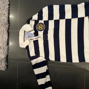 Randig pikétröja Hampton Republic - Säljer en klassisk långärmad pikétröja från Hampton Republic i marinblått och vitt med breda ränder. Tröjan har krage, knäppning framtill och ett stort broderat märke på bröstet. Perfekt för en preppy look!