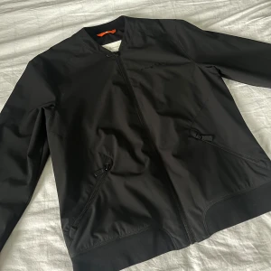 Svart bomberjacka Sail Racing - Svart bomberjacka från Sail Racing med stilren design, hel dragkedja framtill och två snedställda fickor med dragkedja. Jackan har ribbade muddar vid ärmslut och nederkant samt diskret logga på bröstet. Perfekt för en clean och sportig look.