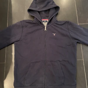 Mörkblå hoodie från GANT med dragkedja - Säljer en mörkblå hoodie från GANT med hel dragkedja och huva. Tröjan har två stora fickor framtill och diskret GANT-logga på bröstet. Tillverkad i mjuk bomull, perfekt för chill dagar. Klassisk och stilren design.