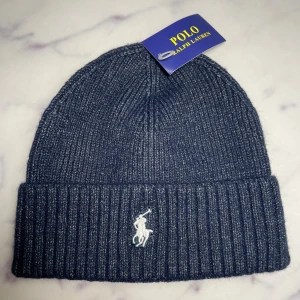Mörkblå ribbstickad mössa Polo Ralph Lauren - Ribbstickad mörkblå mössa från Polo Ralph Lauren med klassisk uppvikt kant och broderad vit logga framtill. Perfekt accessoar för kyliga dagar, tillverkad i mjukt och värmande material. Tveka inte att höra av er om ni har frågor/prisförslag!🙌