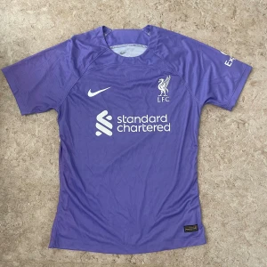 Lila Liverpool FC Nike fotbollströja - Säljer en lila Liverpool FC fotbollströja från Nike med klubbmärke och sponsortryck på bröstet. Tröjan har korta ärmar, rund hals och är tillverkad i ett lätt, ventilerande funktionsmaterial. Perfekt för dig som vill sticka ut på planen eller på läktaren.